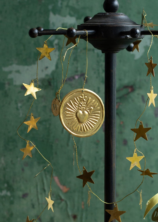 Brass Sacred Heart ornament with sunray design hanging from a dark metal stand, surrounded by a gold star garland against a green textured background.