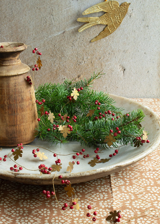 A gold-colored brass garland with oak leaves and red berries displayed on a plate next to a wooden vase and a decorative bird ornament.