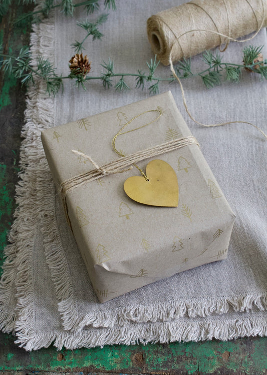 A brass handmade heart ornament placed on a wrapped gift with a beige paper, on a surface with a roll of twine and greenery in the background.