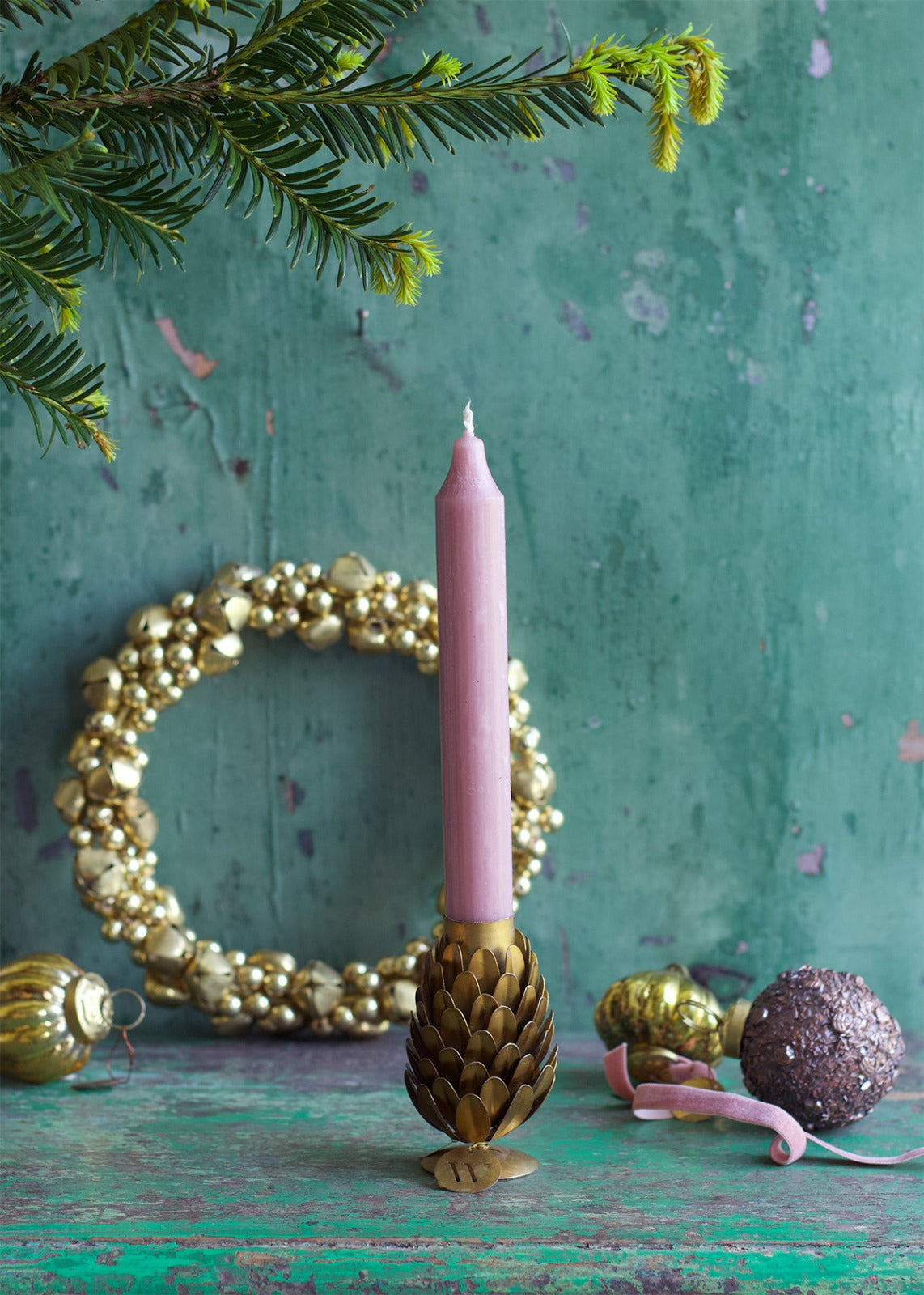 Two brass pinecone-shaped candle holders on a wooden surface with lit candles in them, surrounded by decorative pine cones and a decorative wall hanging in the background.