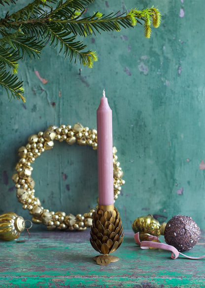 Two brass pinecone-shaped candle holders on a wooden surface with lit candles in them, surrounded by decorative pine cones and a decorative wall hanging in the background.