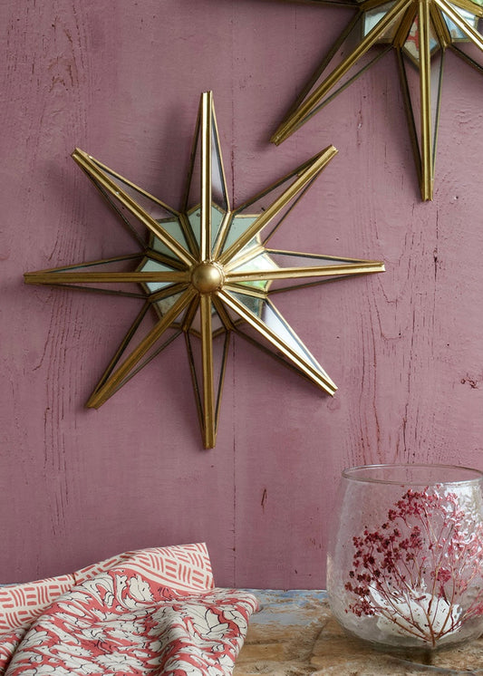 A gold-colored star-shaped mirror hanging on a purple wall, with a glass vase and a red patterned object to the side.