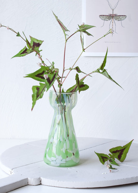 Handmade green and white speckled glass vase with hourglass shape, styled with wild foliage on a white table.

