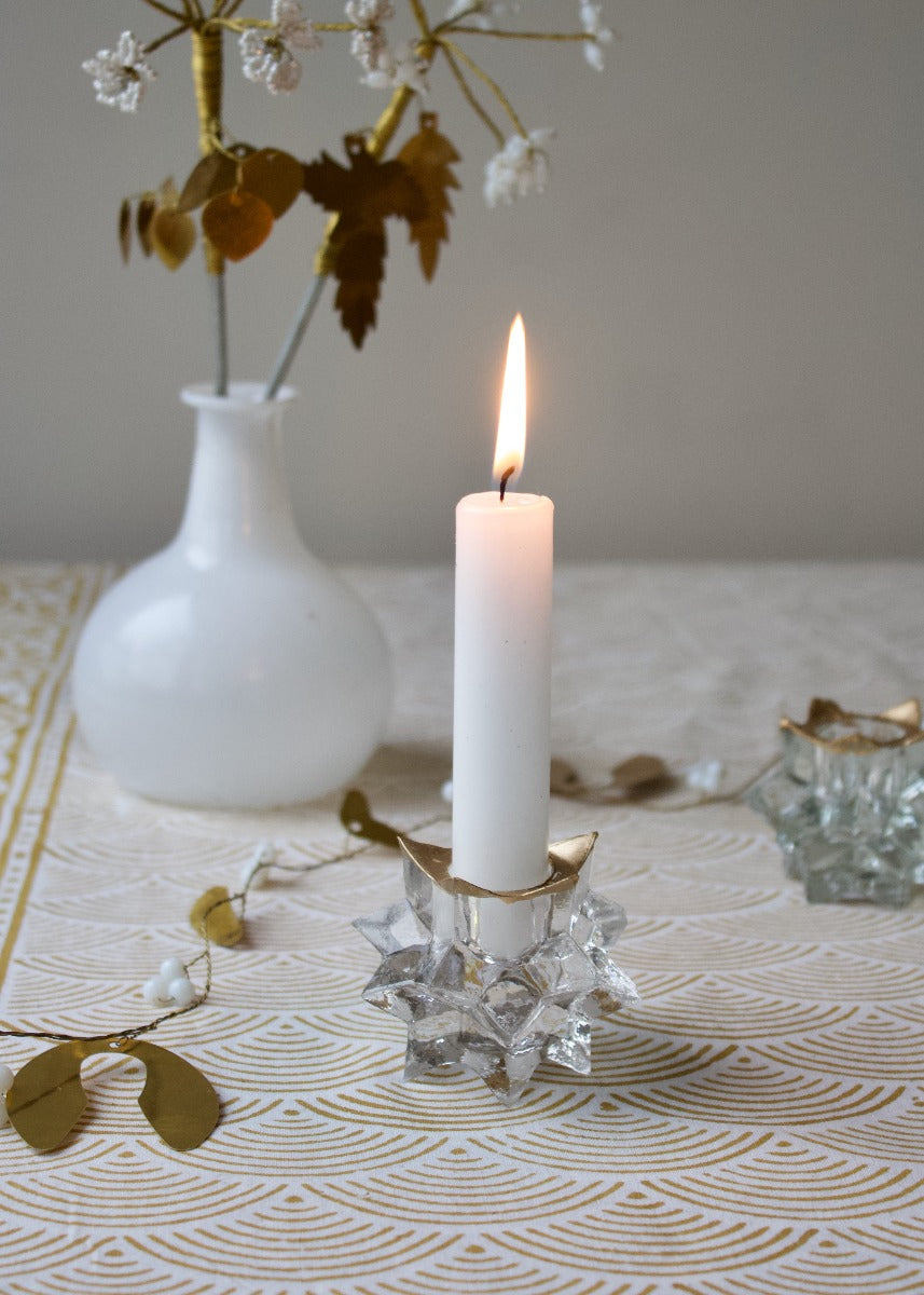 A white candle is lit and placed on a glass candle holder with gold-colored accents, which is placed on a table with a decorative patterned tablecloth, alongside a vase with dried flowers and other decorative items.