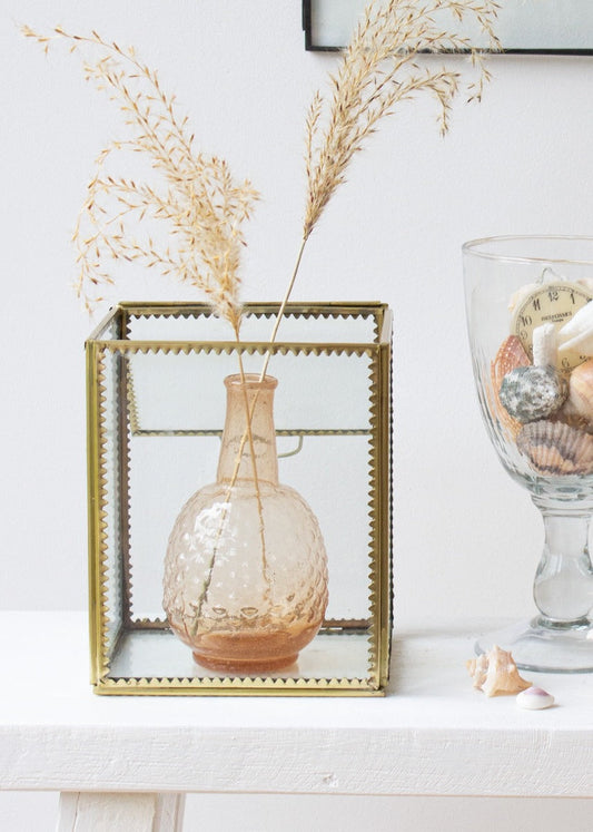 A small amber glass vase with dried grasses displayed inside a brass and glass display box on a white table, next to a tall glass filled with shells and vintage items.