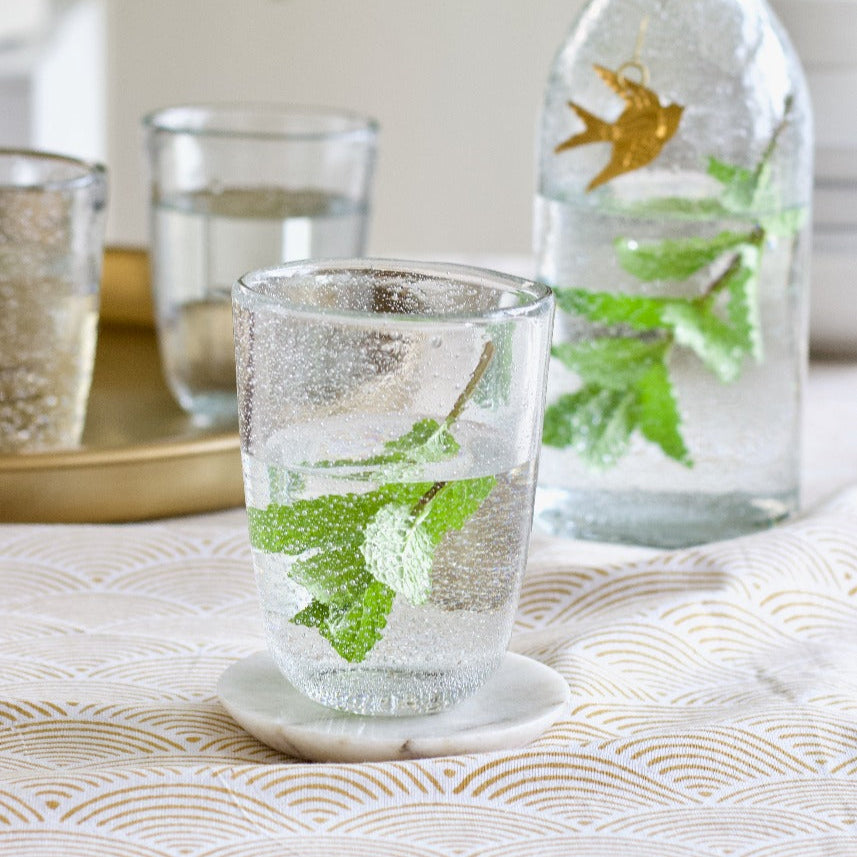 Clear glass with water and mint leaves on a textured surface with a pitcher in the background.