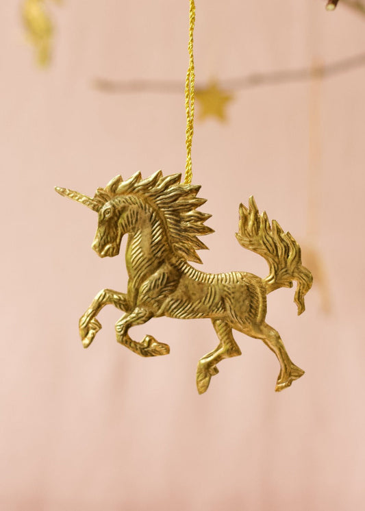 Brass unicorn ornament with a flowing mane and tail, hanging from a golden thread against a soft pink background.