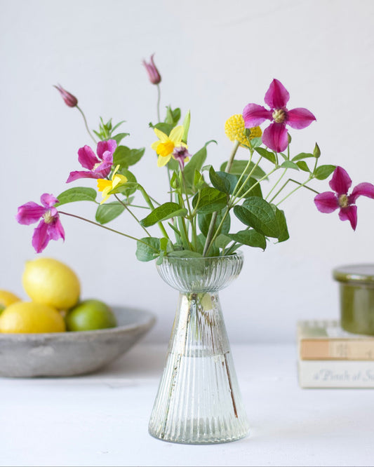 Clear glass vase with colorful flowers on a white surface with lemons and a book in the background.