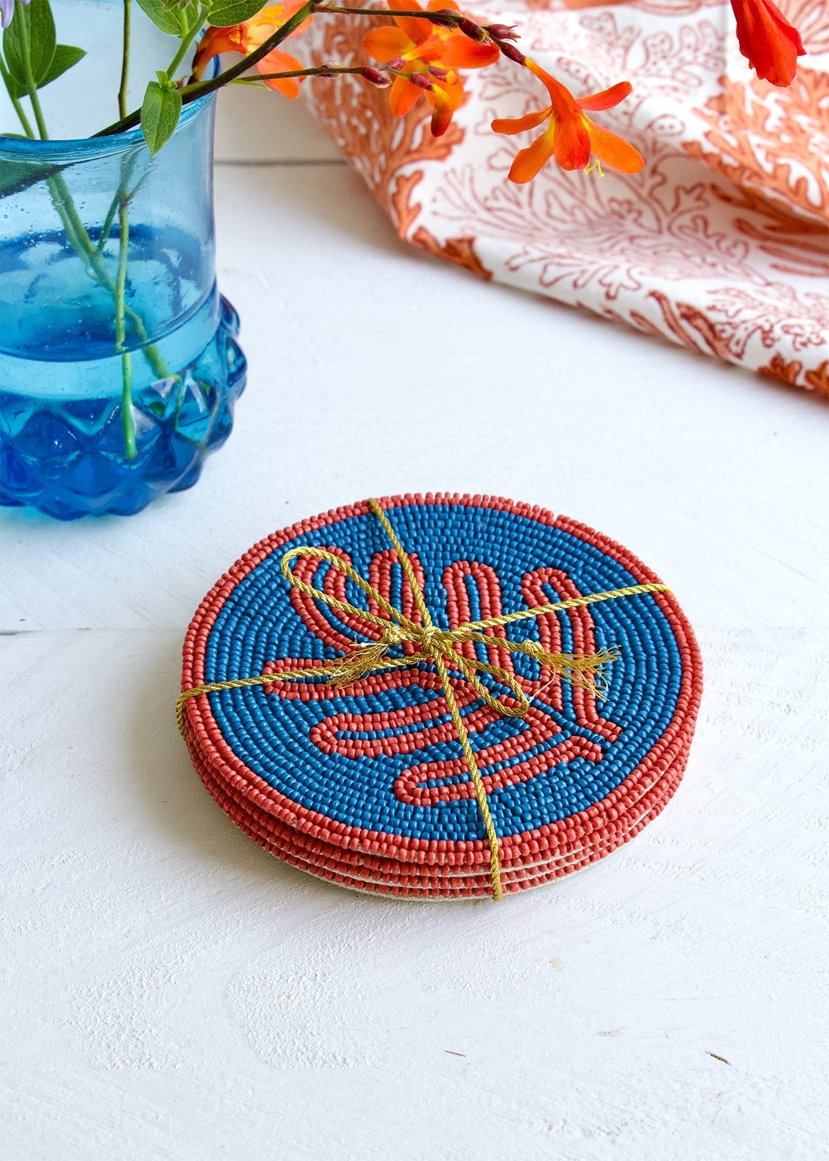 Set of beaded coasters with a lobster design on a white surface, next to a blue vase with flowers and a patterned cloth.