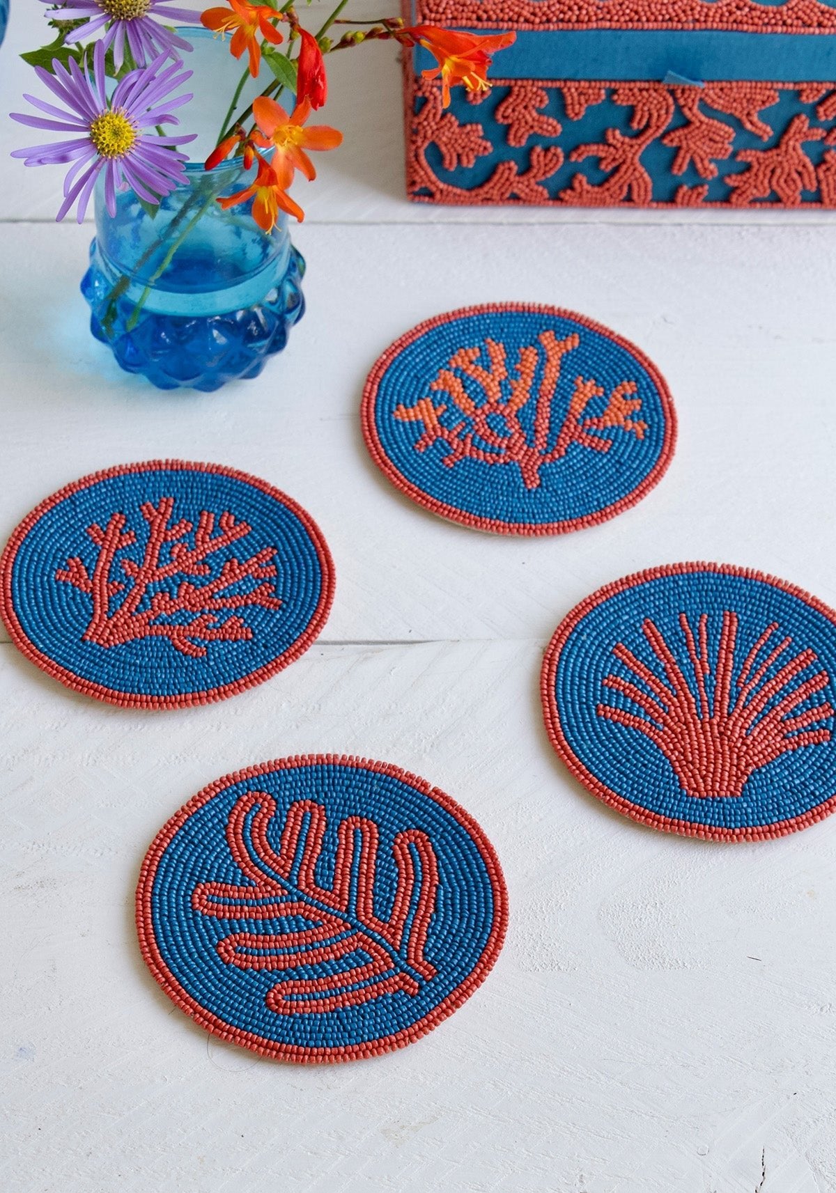 Set of four blue and orange embroidered coasters on a white surface with a vase of flowers.