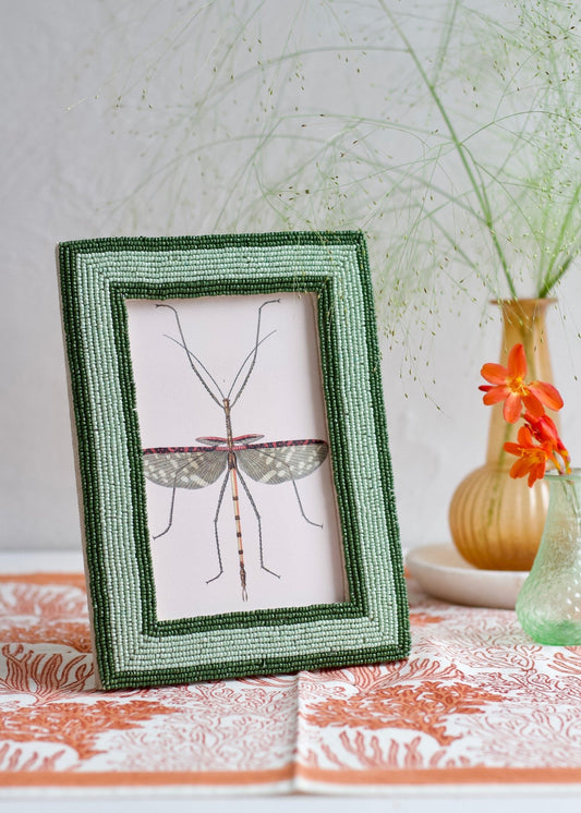 Framed artwork of a dragonfly on a table with a vase and flowers in the background