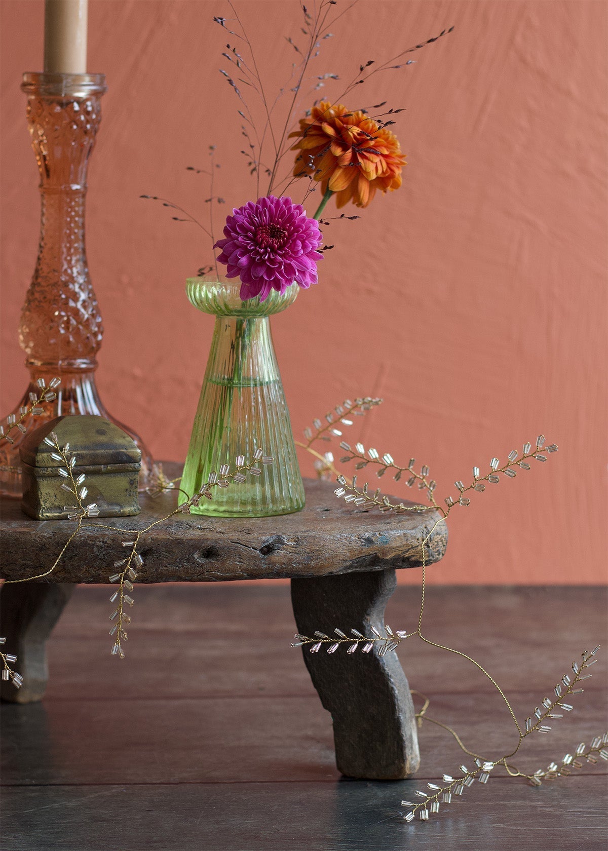 Decorative still life with a green vase, pink candle, and flowers on a wooden stool against a pinkish-brown background.