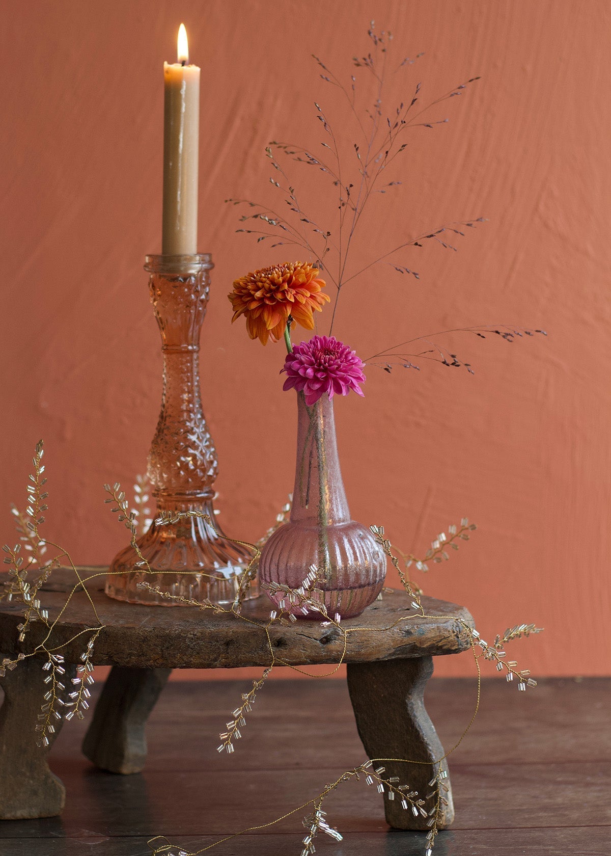 Decorative setup with a candle, vase with flowers, and decorative elements on a wooden surface against a red-brown background.