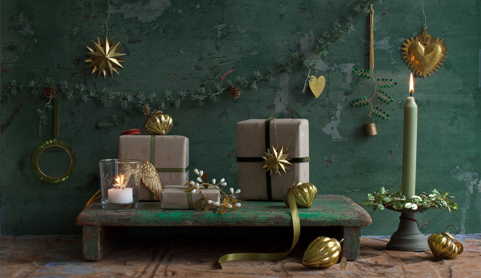 Festive Christmas scene with wrapped gifts, golden ornaments, a green candle in a wreath-shaped holder, a burning tealight, and decorative garlands on a rustic green background.