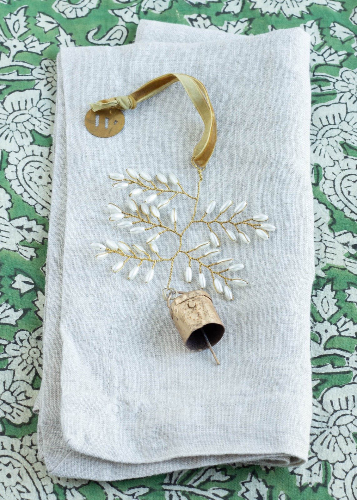 Brass snowflake ornament with white beads and a small rustic bell, displayed on a folded linen napkin with a green floral background.