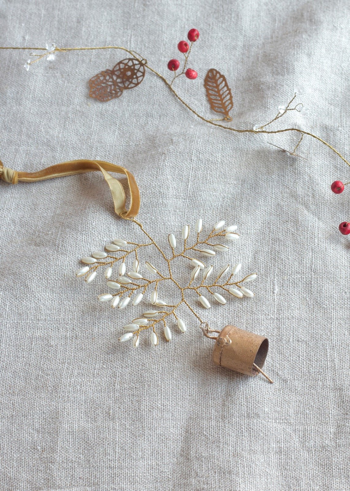 Handmade brass snowflake ornament with pearl-like beads and a small bell, resting on natural linen with a decorative garland of golden leaves and red berries.