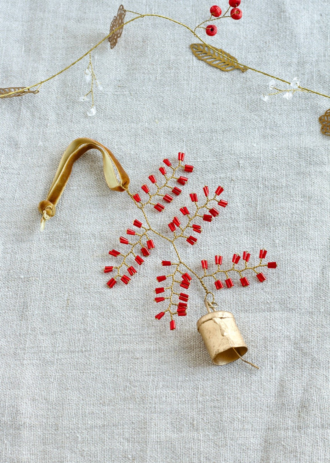 Flatlay of a handmade ornament shaped like a delicate leaf, crafted from red glass beads and gold wire, with a small brass bell and a velvet ribbon, placed on a linen background.