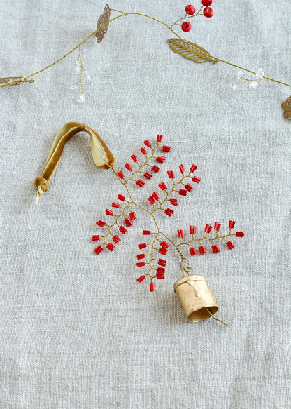 Flatlay of a handmade ornament shaped like a delicate leaf, crafted from red glass beads and gold wire, with a small brass bell and a velvet ribbon, placed on a linen background.