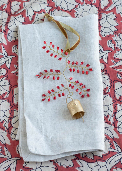 Handmade ornament with red glass beads and brass bell, displayed on folded linen fabric with a floral patterned background.