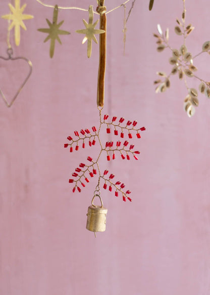 Handmade brass snowflake ornament with red glass beads and a small bell, hanging from a golden velvet ribbon against a soft pink background.