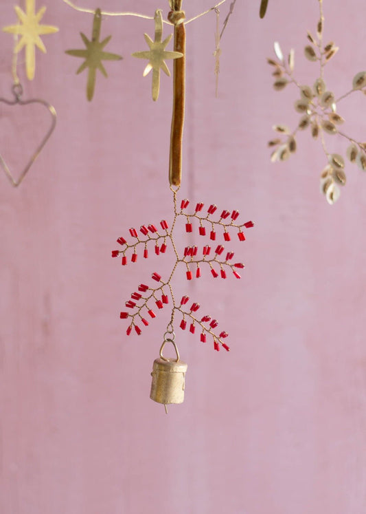 Handmade brass snowflake ornament with red glass beads and a small bell, hanging from a golden velvet ribbon against a soft pink background.