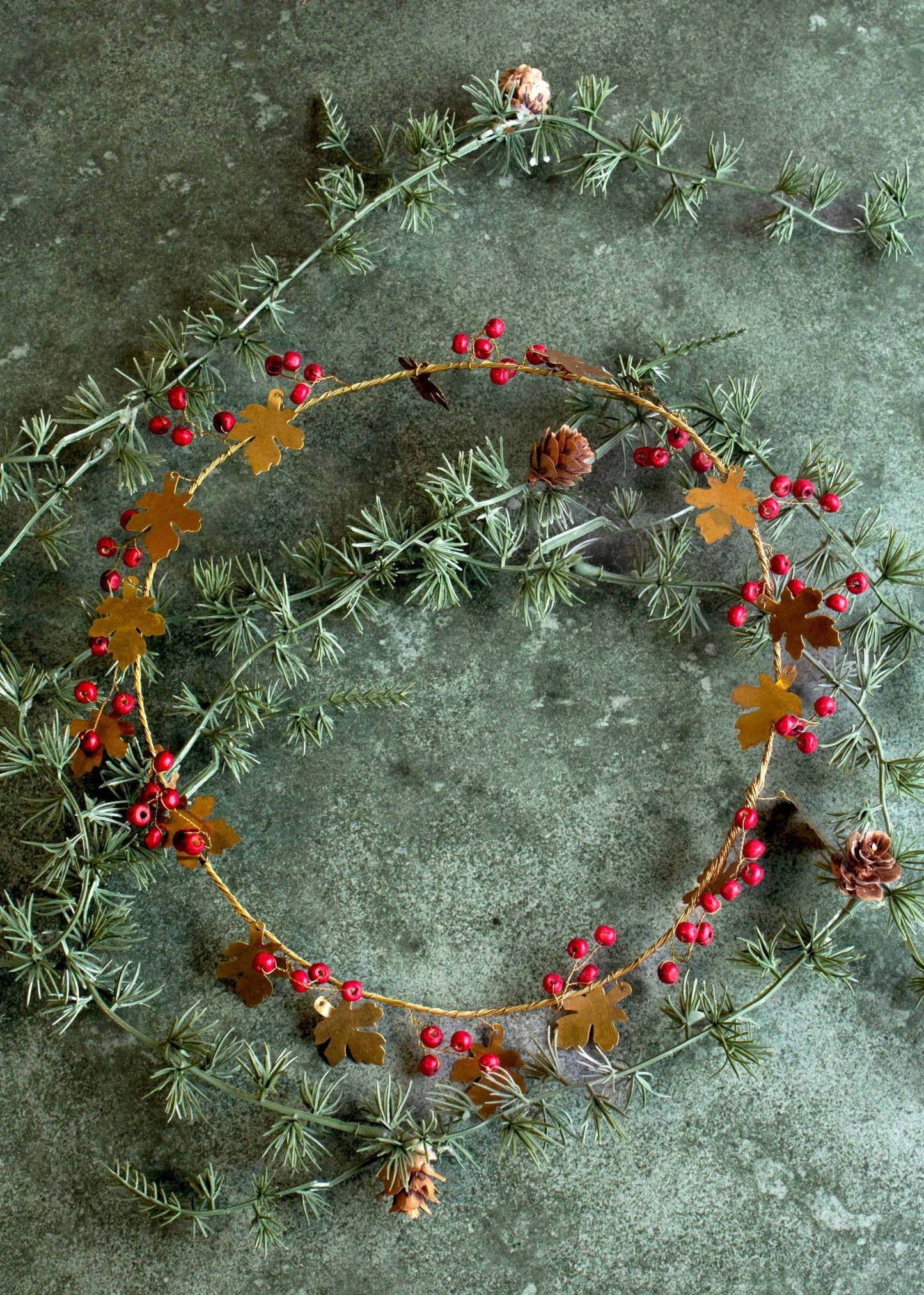 A decorative handmade brass wreath with oak leaves and red berries displayed on a dark textured surface.