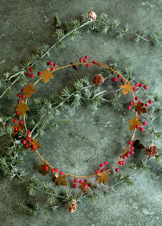 A decorative handmade brass wreath with oak leaves and red berries displayed on a dark textured surface.