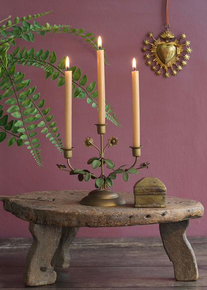 Brass candle holder with three taper candles and leaf details on a rustic wooden table against a pink wall.