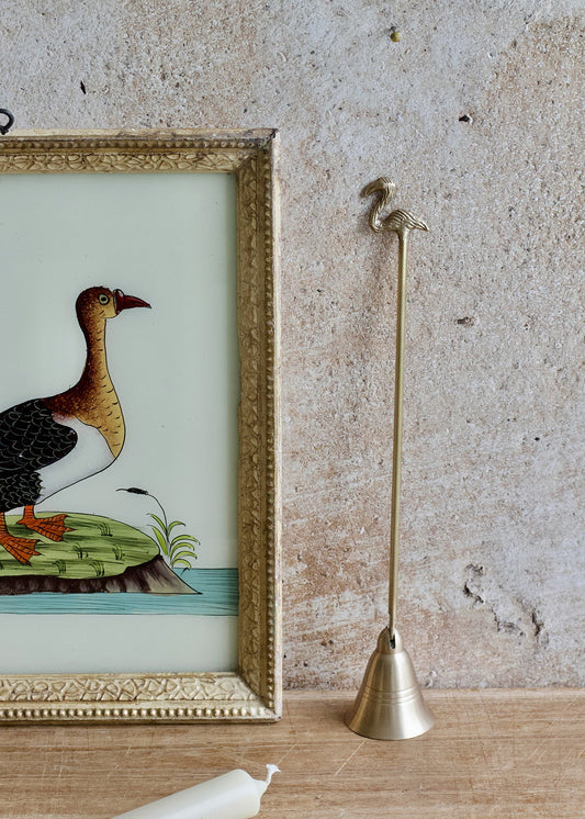 Brass candle snuffer next to a framed illustration of a duck on a textured wall.