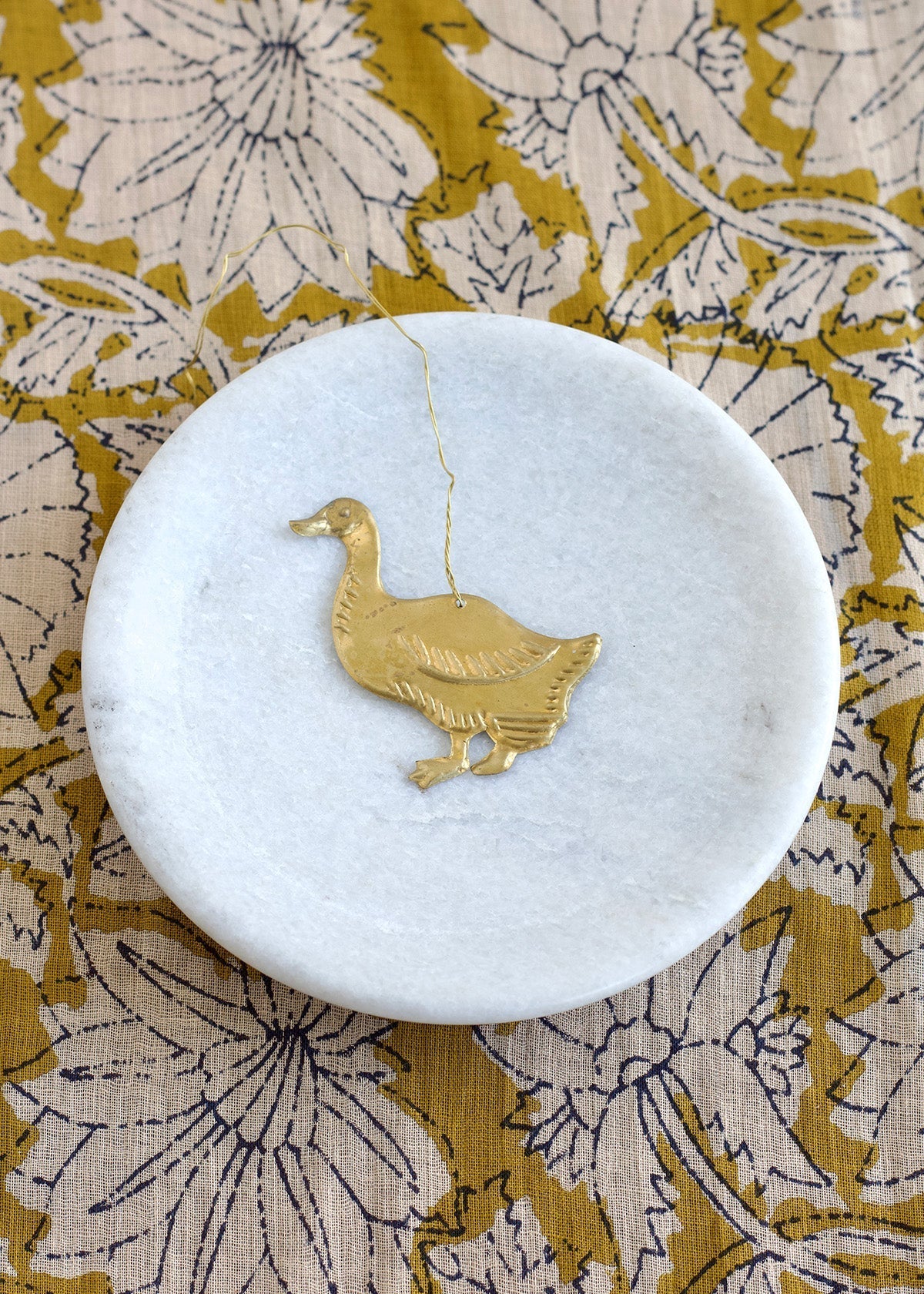 Brass duck ornament placed on a round white marble dish, with a floral mustard-yellow fabric underneath.

