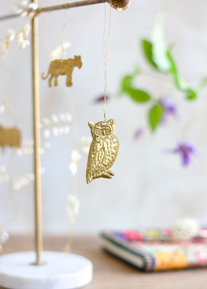 Brass owl ornament hanging from a wire, with soft background elements including blurred animal shapes and flowers.