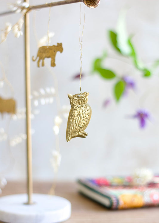 Brass owl ornament hanging from a wire, with soft background elements including blurred animal shapes and flowers.