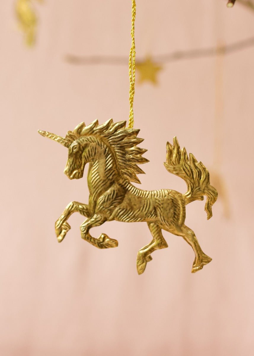 Brass unicorn ornament with a flowing mane and tail, hanging from a golden thread against a soft pink background.