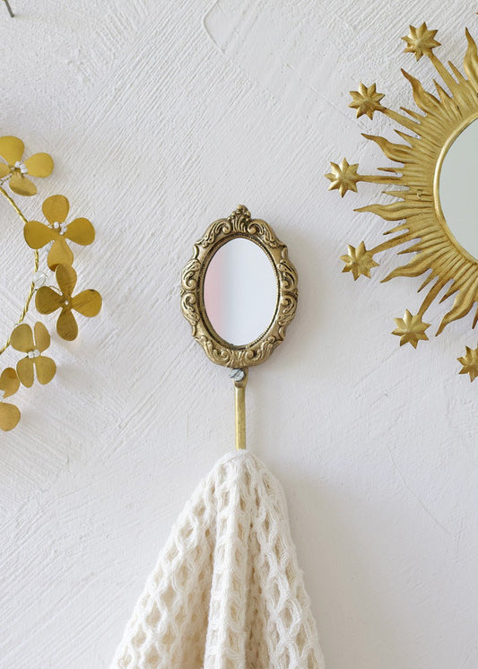 Decorative wall hook with a small mirror and white towel on a white wall with gold accents.