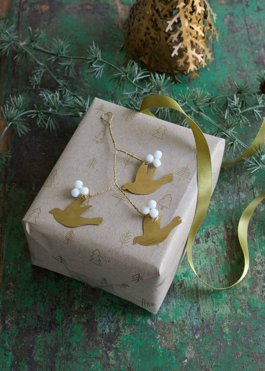 Golden brass ornament with three birds and white mistletoe bead details, displayed on a wrapped gift.