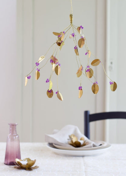 Small decorative brass bunch with oval leaves and purple beads, hanging above a table set with a soft linen napkin and vintage pink vase.