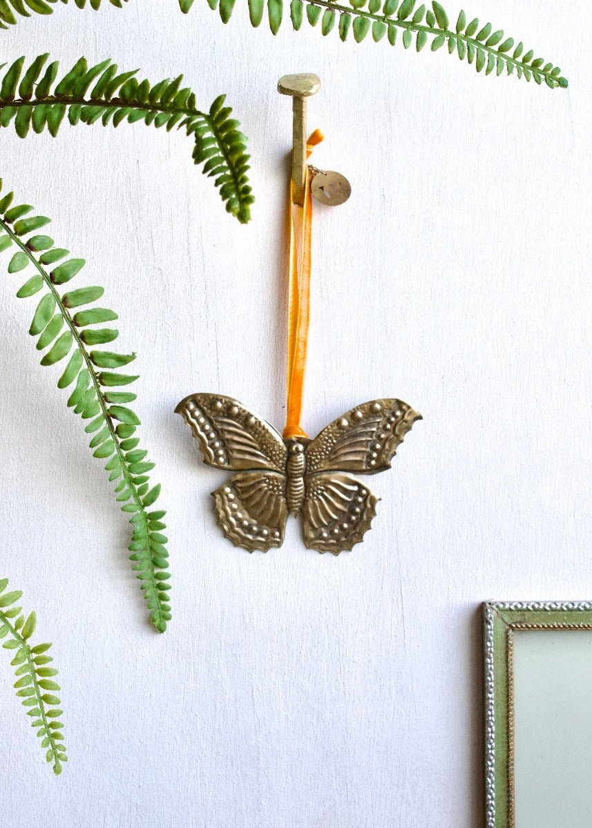 Zinc butterfly ornament with orange ribbon, hanging on a white wall surrounded by green fern leaves.