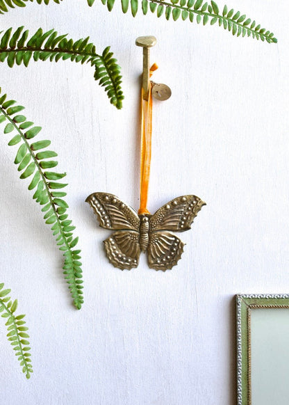 Zinc butterfly ornament with orange ribbon, hanging on a white wall surrounded by green fern leaves.