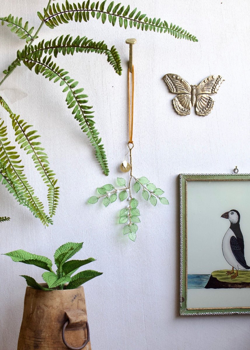 Decorative green leaf branch ornament hanging from an orange ribbon on a white wall, surrounded by fern leaves, a zinc butterfly, and a framed puffin illustration.
