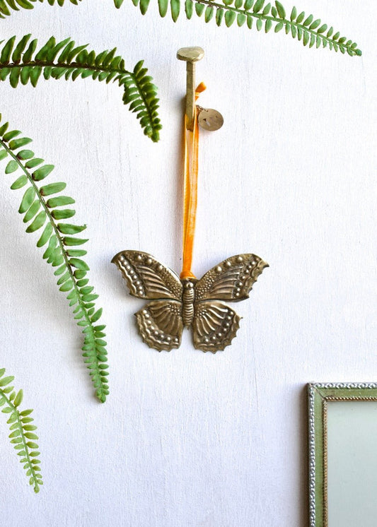 Zinc butterfly ornament with orange ribbon, hanging on a white wall surrounded by green fern leaves.