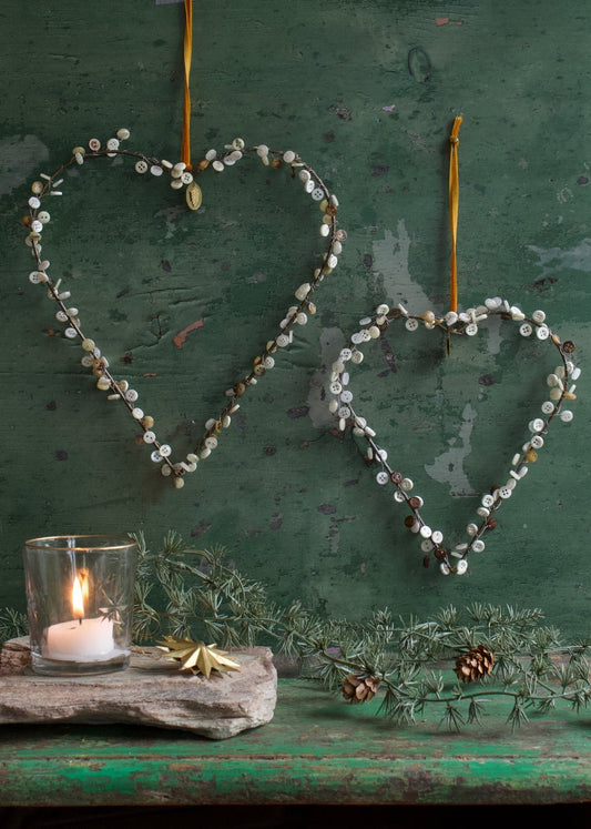 Two heart-shaped wire ornaments with vintage buttons, hanging from golden ribbon against a rustic green background, with a lit candle and pine branch below.