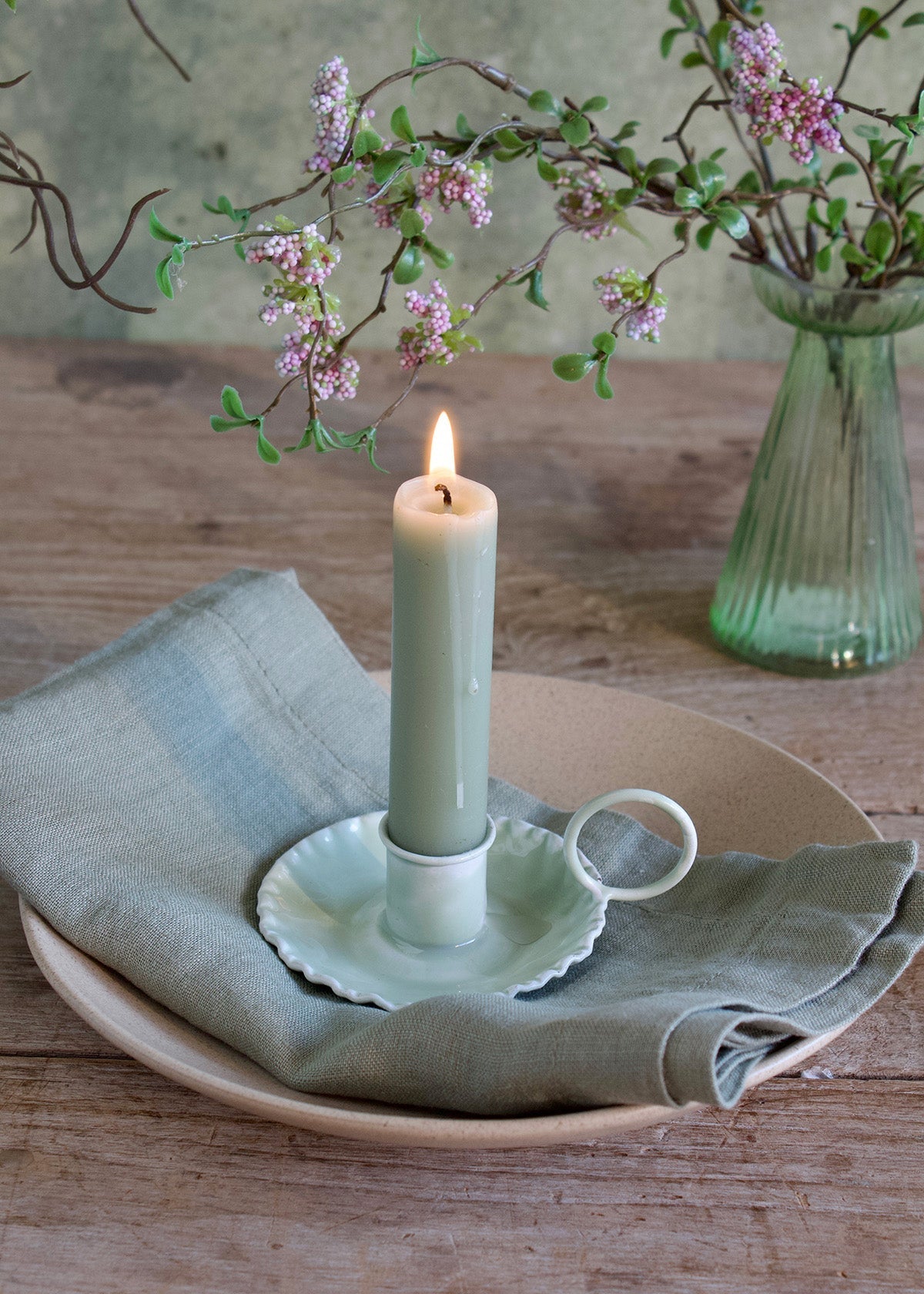 Candle in a decorative holder on a wooden table with a vase of flowers in the background