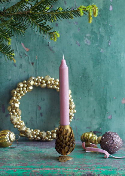 Two brass pinecone-shaped candle holders on a wooden surface with lit candles in them, surrounded by decorative pine cones and a decorative wall hanging in the background.