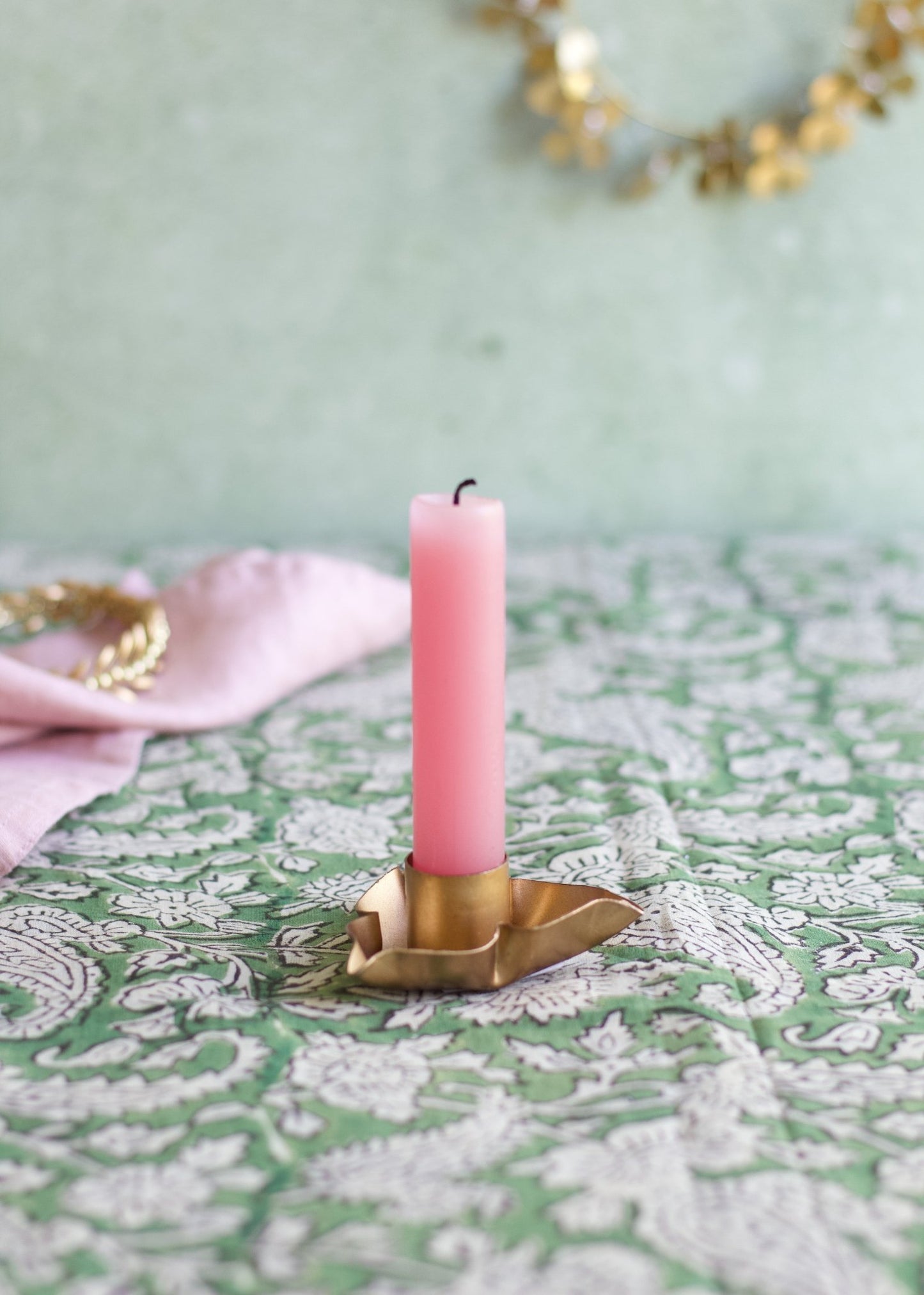 Pink candle in a brass holder on a decorative tablecloth with a light green background