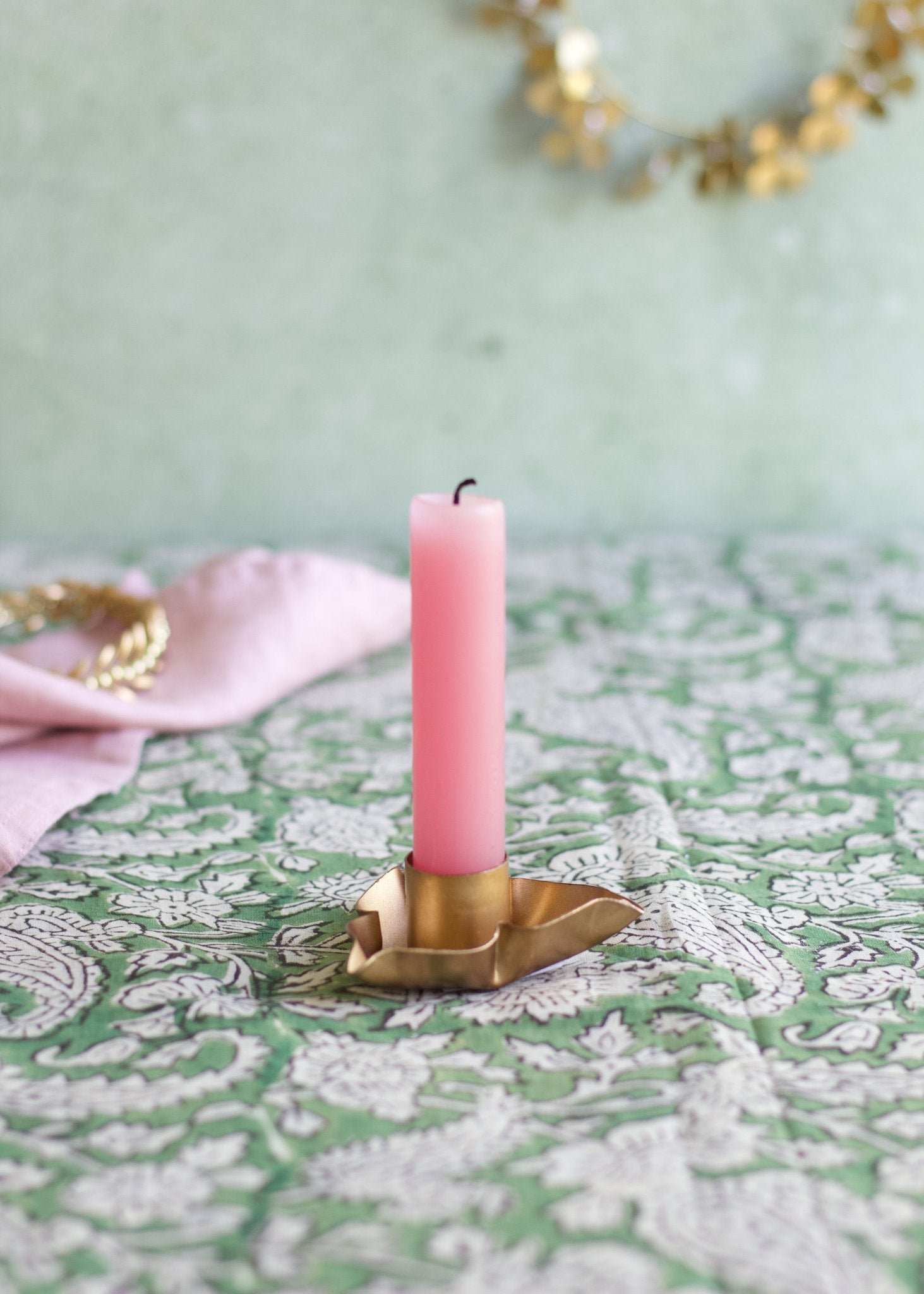 Pink candle in a brass holder on a decorative tablecloth with a light green background