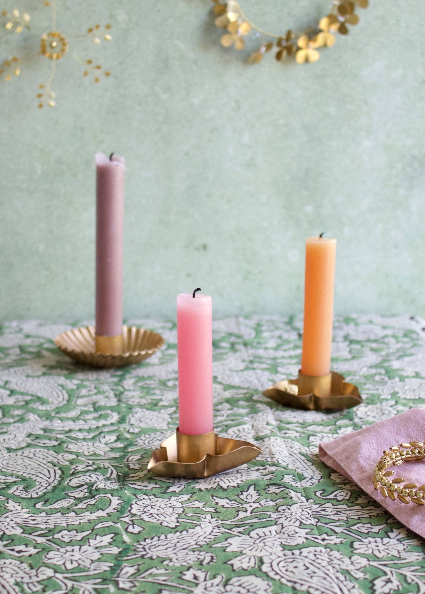 Three candles in brass candle holders on a patterned tablecloth with a decorative gold garland in the background.