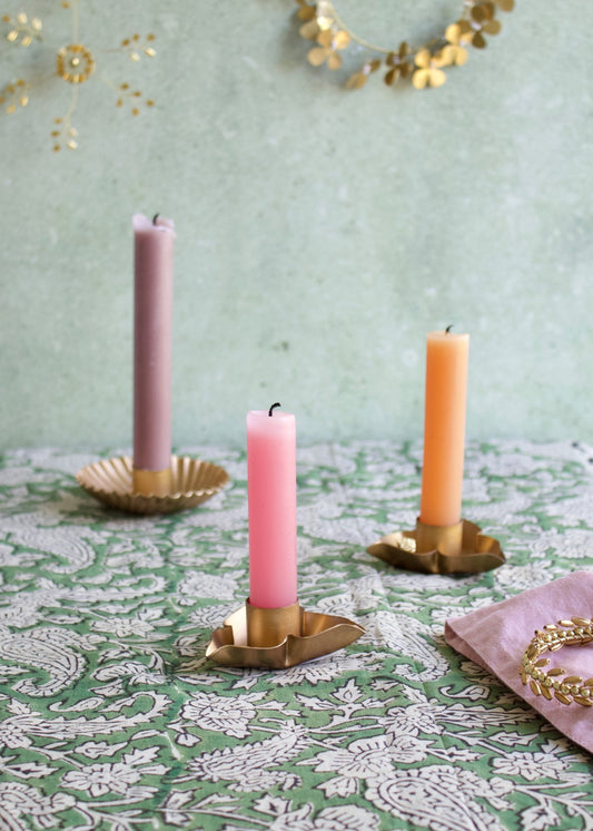 Three candles in brass candle holders on a patterned tablecloth with a decorative gold garland in the background.