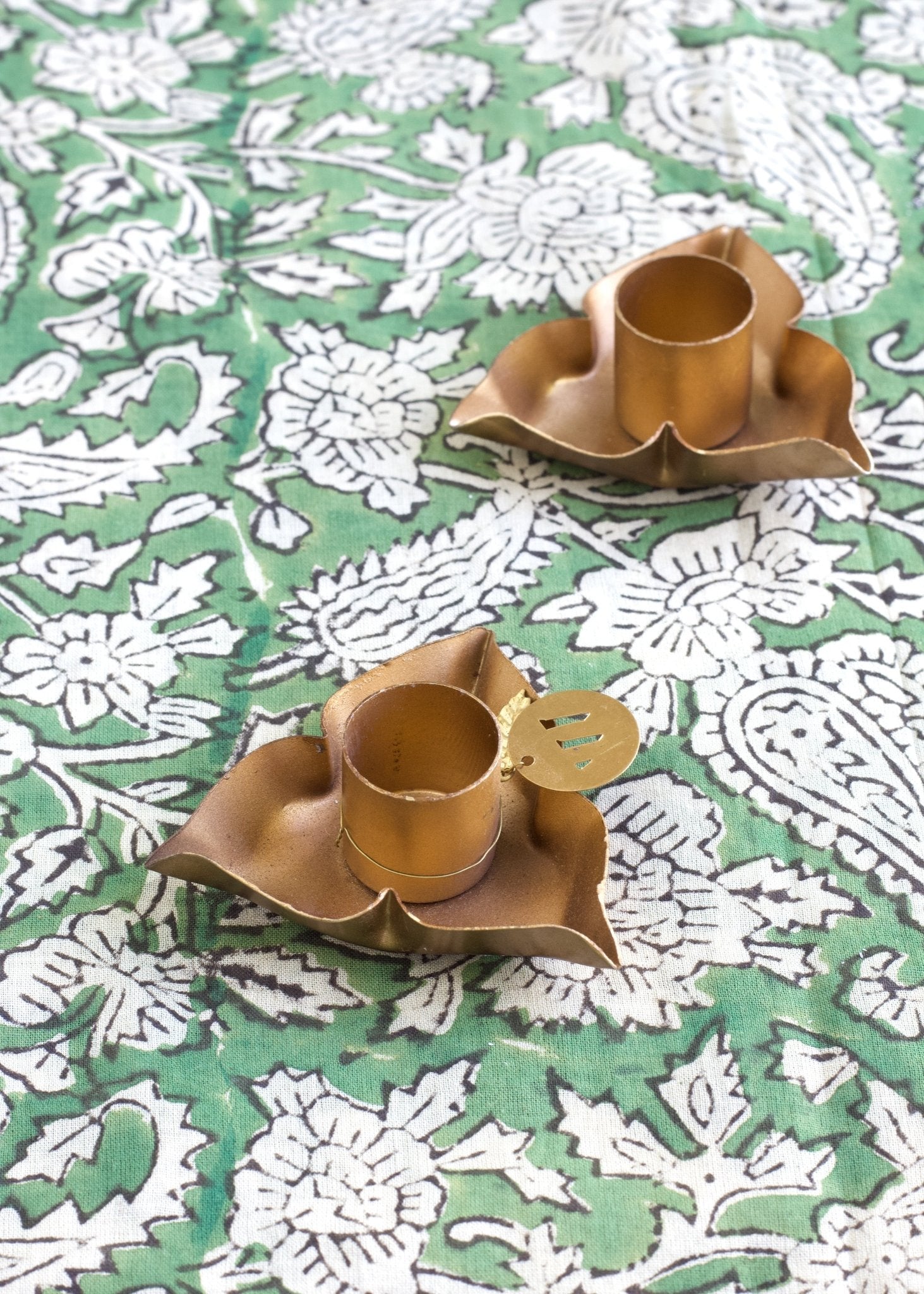 Brass candleholders with a petal shape inspired by the Oxalis flower, placed on a green and white floral block-printed fabric.
