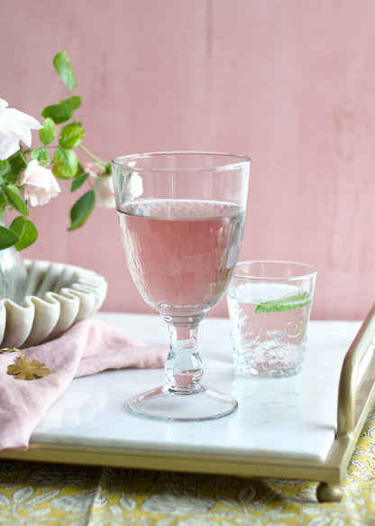 A clear handmade wine glass filled with a pink liquid, placed on a table with a floral patterned napkin and a small vase of flowers in the background.