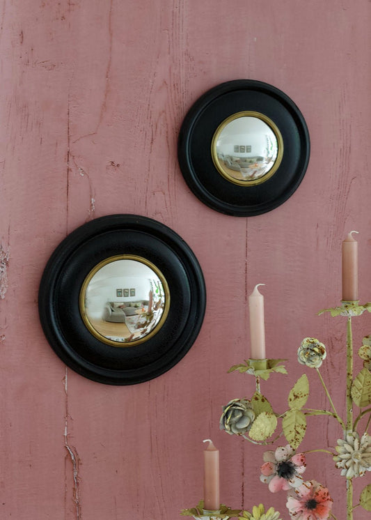 Two round convex mirrors with black wooden frames and brass inner rims, hanging on a vintage pink wooden wall next to a floral candleholder.
